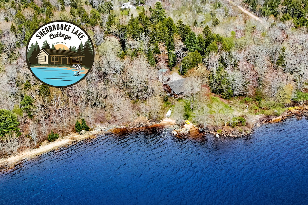 An aerial view captures the cottage nestled among trees along the serene Sherbrooke Lake shoreline. The clear blue water gently laps against the sandy beach, where a small boat is visible, all framed by lush greenery in a tranquil setting.
