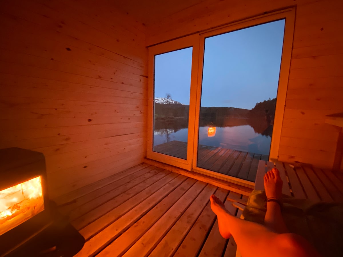 A warm, inviting interior of a cabin features wooden walls and a large glass door overlooking a serene body of water. A small wood burner sits in one corner, casting a soft glow. The floor is wooden, creating a cozy atmosphere.