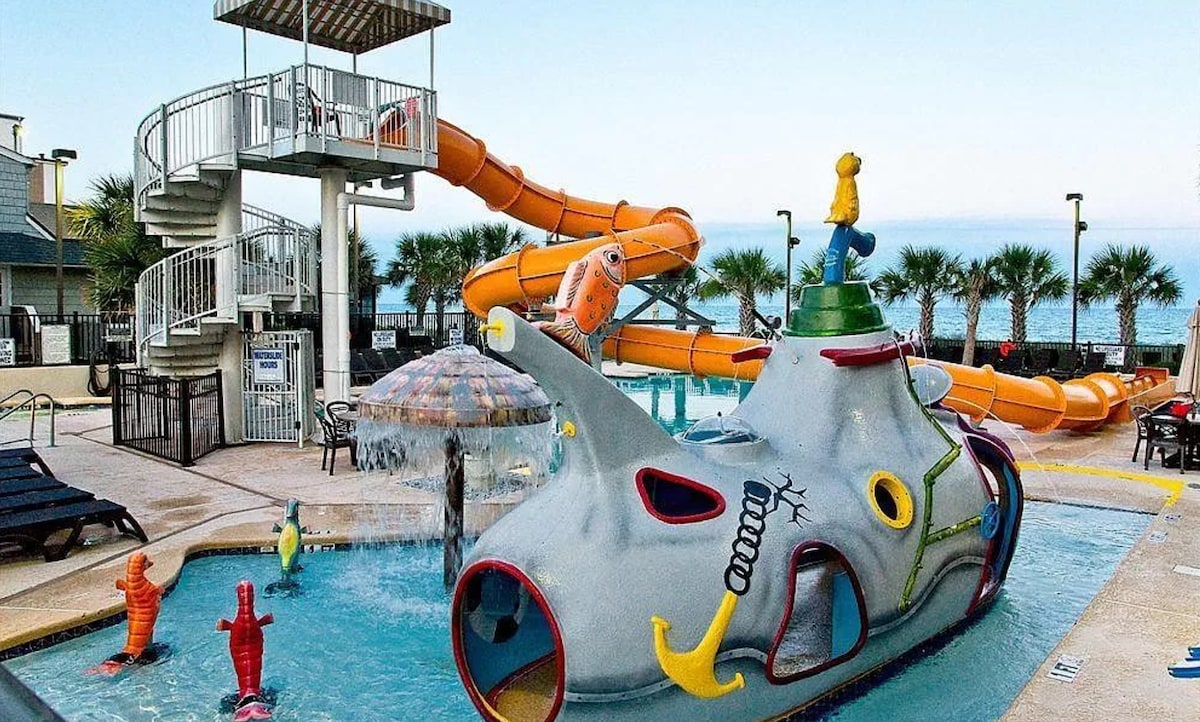 A vibrant children's waterpark is depicted, featuring a colorful creature-themed play structure with slides and water features. A winding orange slide and a spiral slide provide fun options, while palm trees and a distant ocean view add to the outdoor environment.