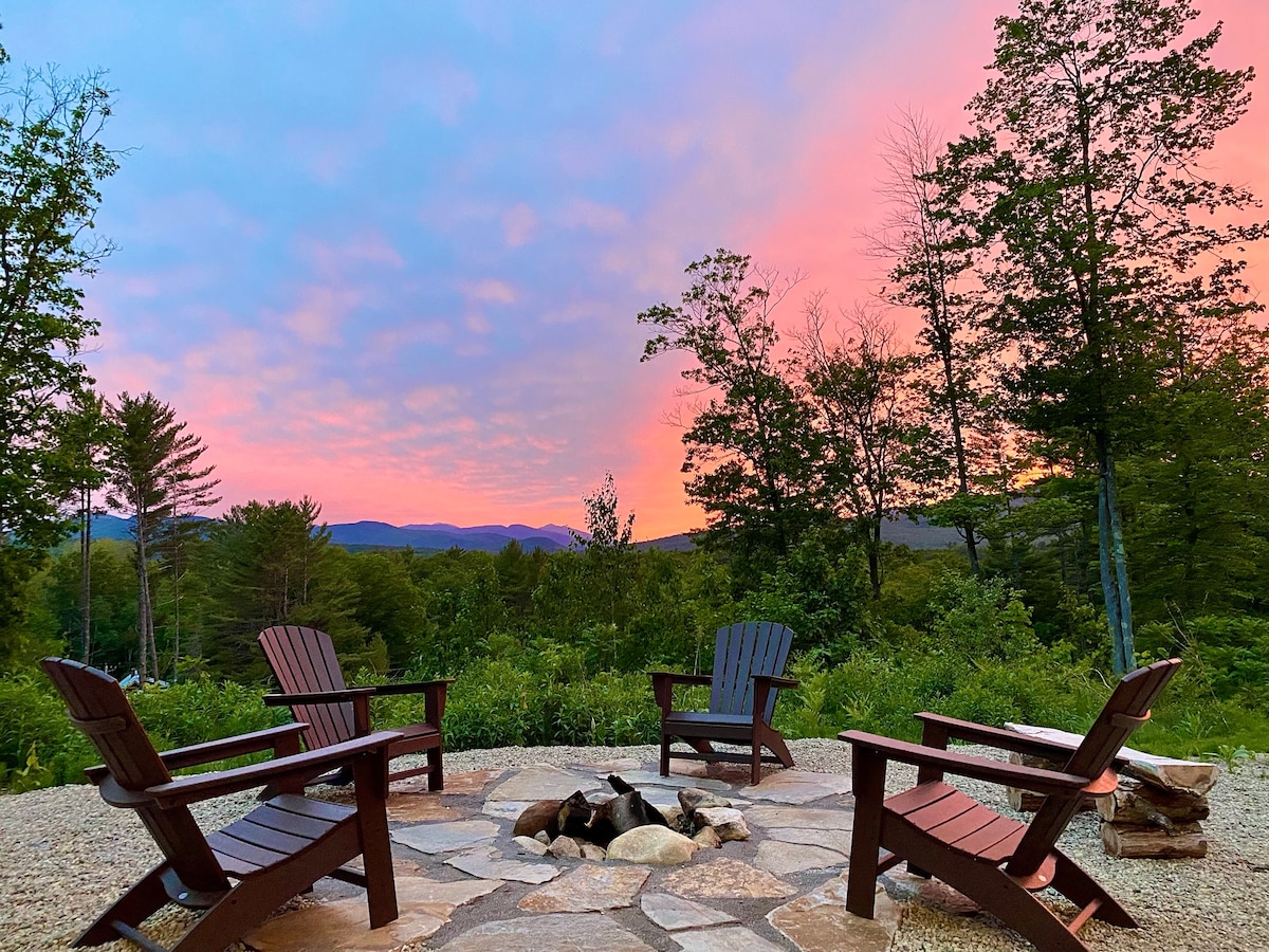 A cozy patio space features four adirondack chairs arranged around a stone fire pit, surrounded by greenery. A scenic view of rolling hills and a colorful sunset sky enhances the outdoor ambiance.