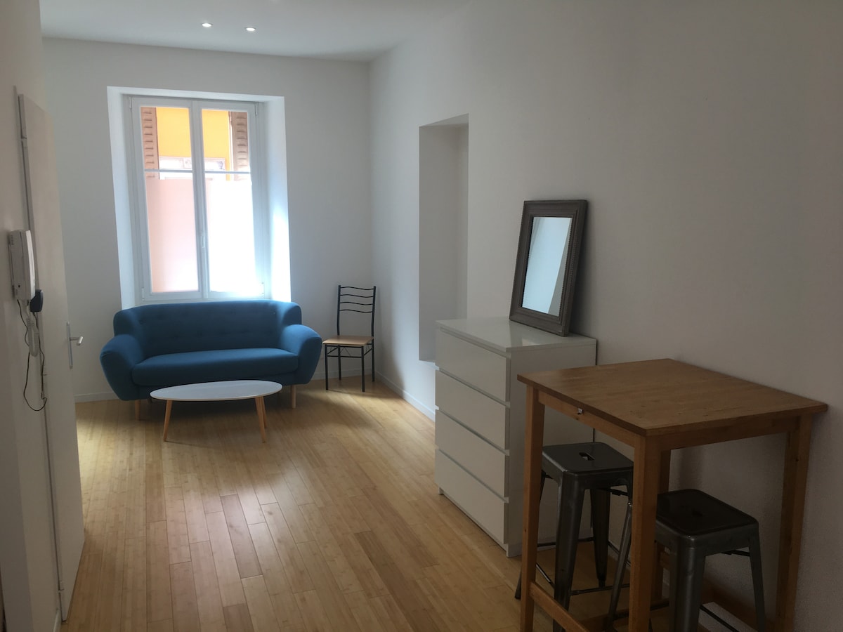 A bright and open living space is showcased, featuring a cozy blue sofa and a round coffee table. A small dining area is presented with a wooden table and metal stools. Natural light floods through a large window, highlighting the wooden flooring and minimalist décor.
