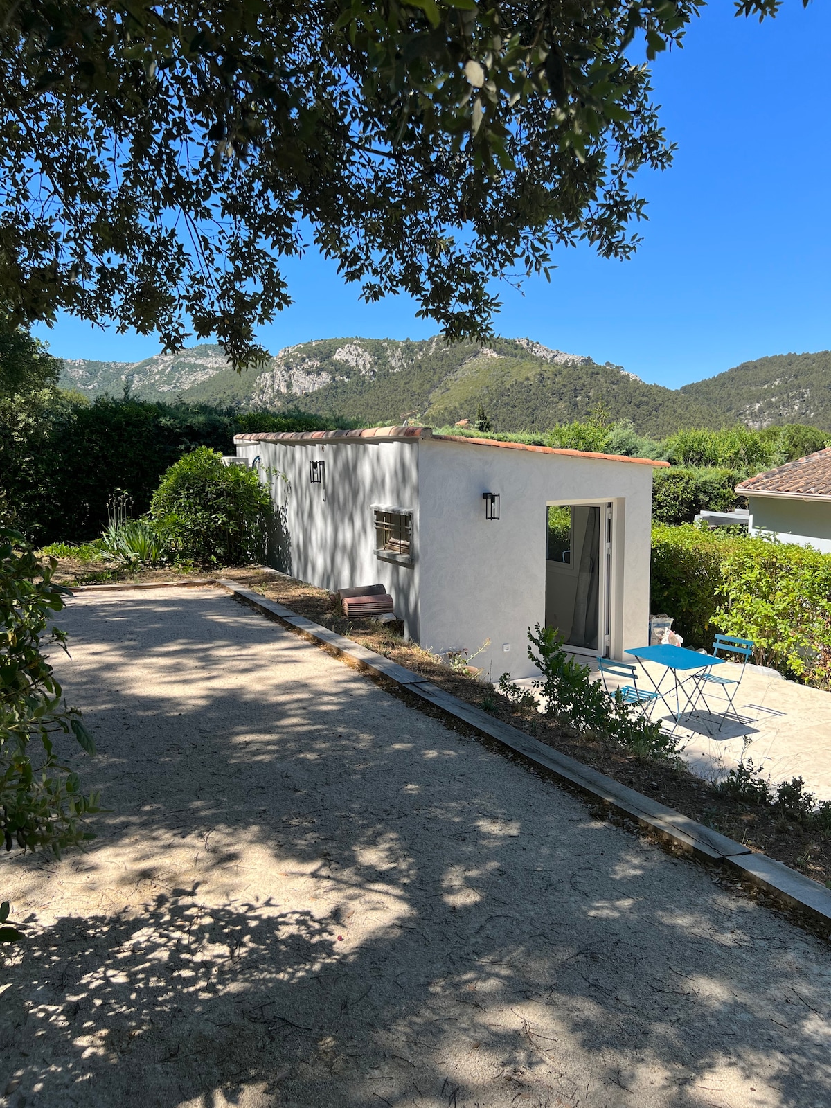 A quaint exterior of the recently renovated dependency is visible, set against a backdrop of the Sainte Baume mountains. A pathway leads to the entrance, flanked by greenery. A small table and chairs are placed outside, inviting a relaxing moment in the serene environment.