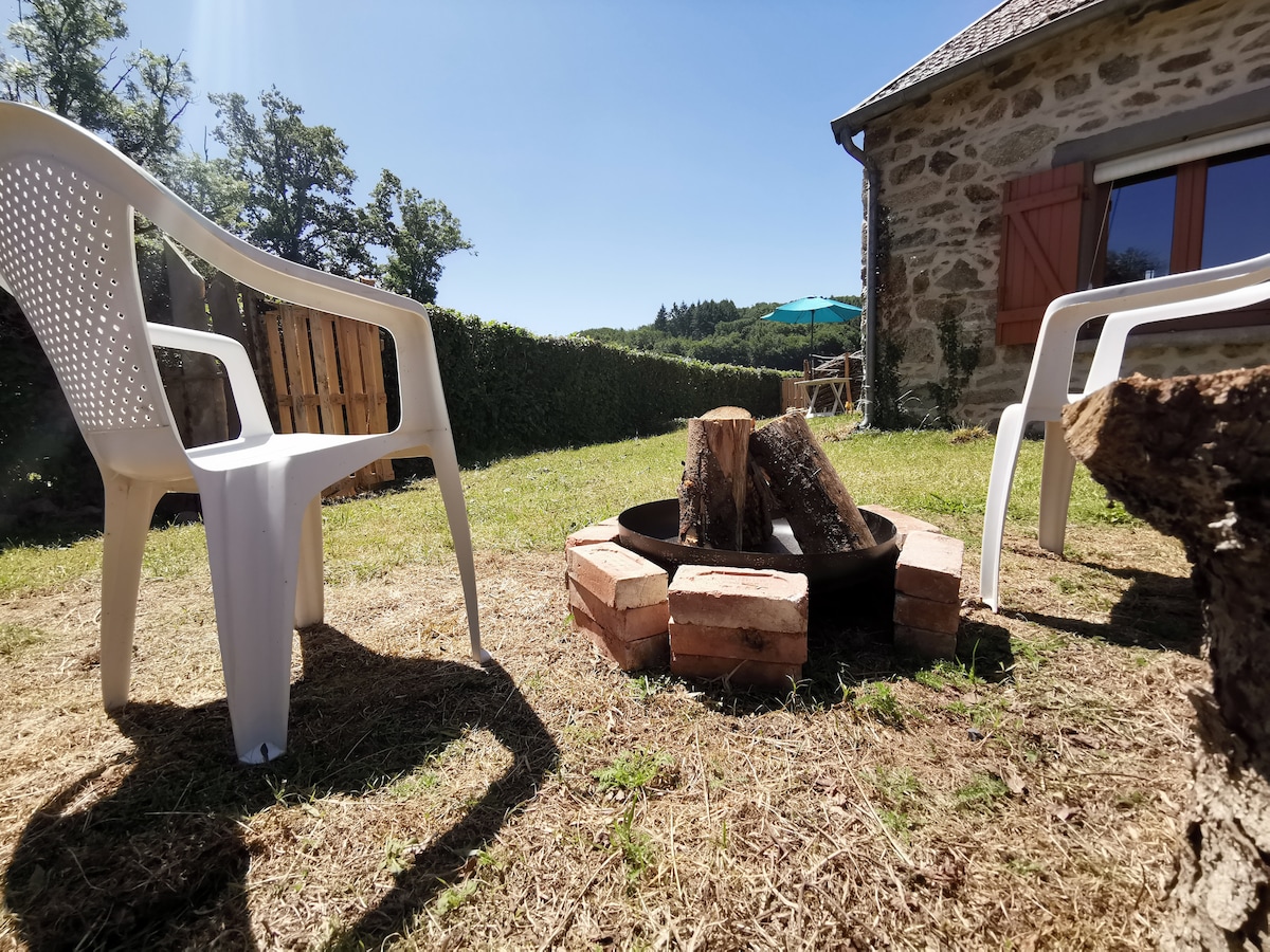 An outdoor space features a circular fire pit surrounded by rustic bricks, with logs placed in the center. Two white plastic chairs are positioned nearby, providing seating for guests. Lush greenery is visible in the background, complementing the serene atmosphere.