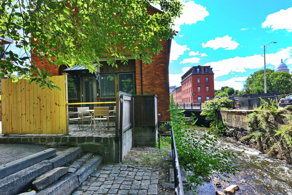 A private outdoor patio is bordered by a wooden fence, featuring seating for four. The view includes the tranquil Moshassuck River, with greenery along the bank. In the background, a red brick building and a historic structure can be seen under a bright blue sky.