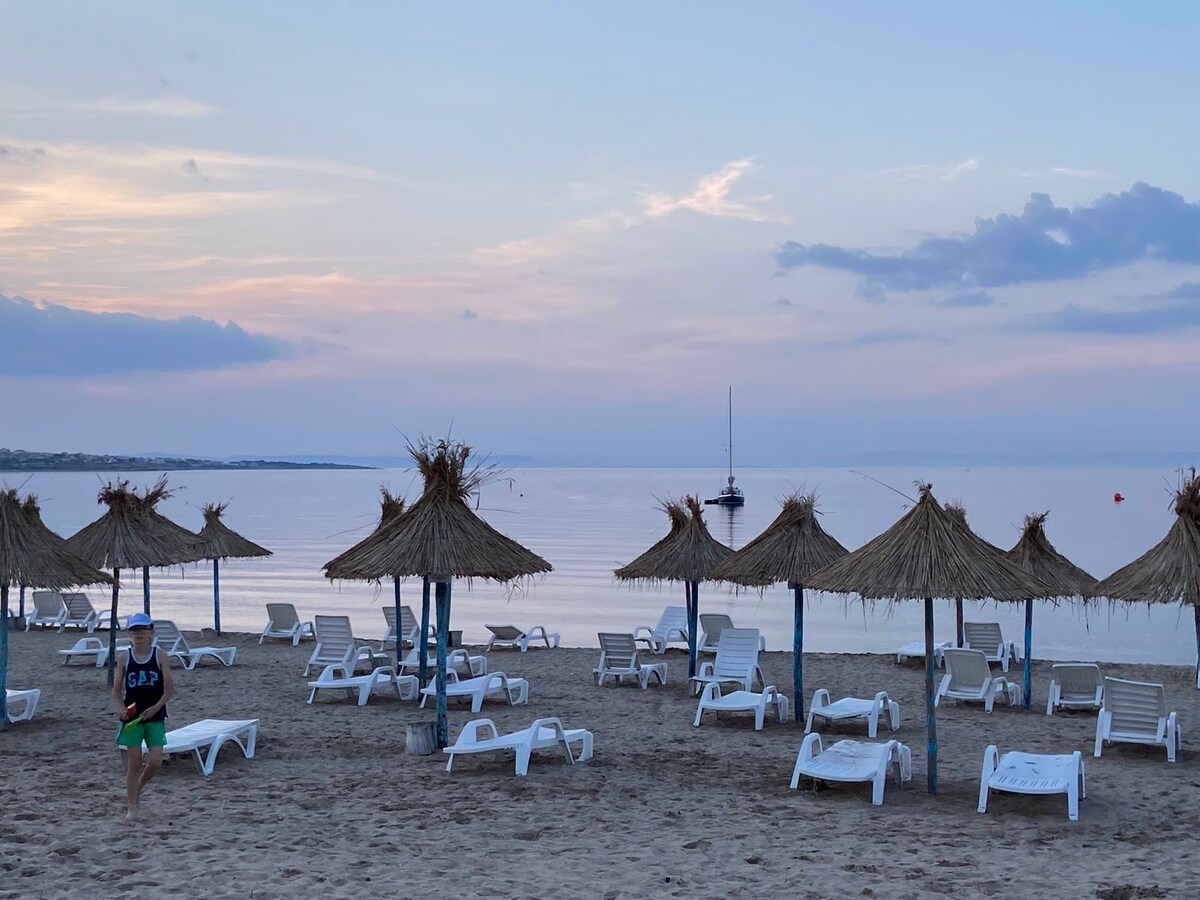 The beach scene features several thatched umbrellas providing shade over white lounge chairs arranged on the sandy shore. A calm sea reflects the pastel hues of the sunset, with a sailboat anchored in the distance under a sky adorned with soft clouds.