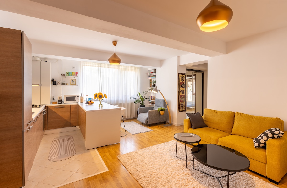 An open living space features a contemporary kitchen with wooden cabinetry and a large island, complemented by a bright living area with a yellow sofa. Sunlight floods through a large window, illuminating a cozy reading nook and modern decor elements.