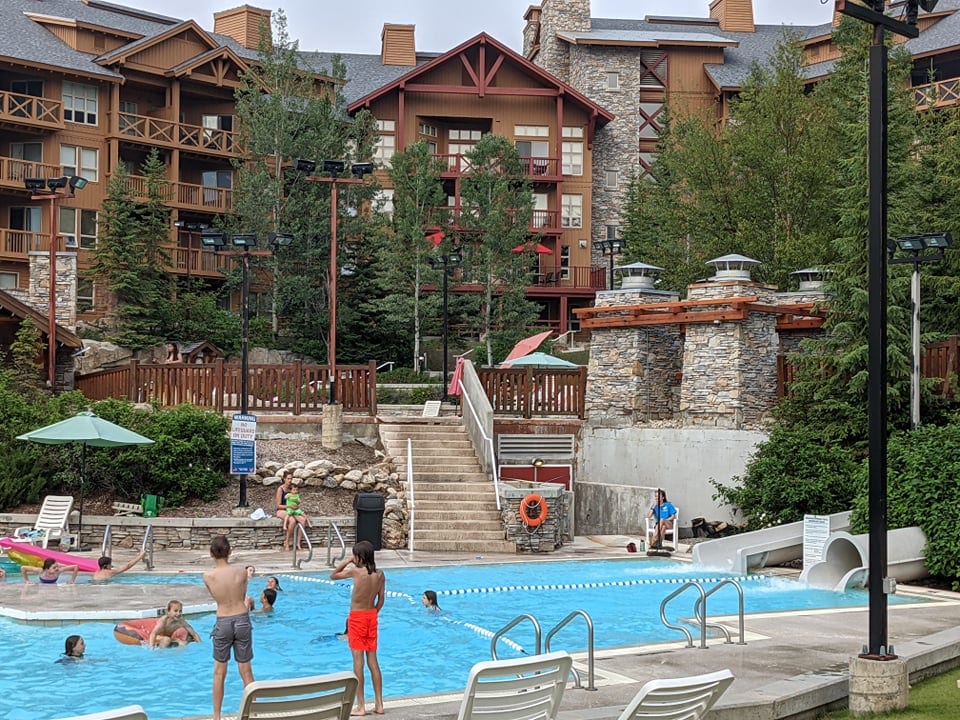 A spacious outdoor pool area is surrounded by lodge-style buildings and lush greenery. Guests are seen enjoying the water, with a range of poolside seating and umbrellas available. A set of stairs leads down from the pool to the grassy area, enhancing accessibility.