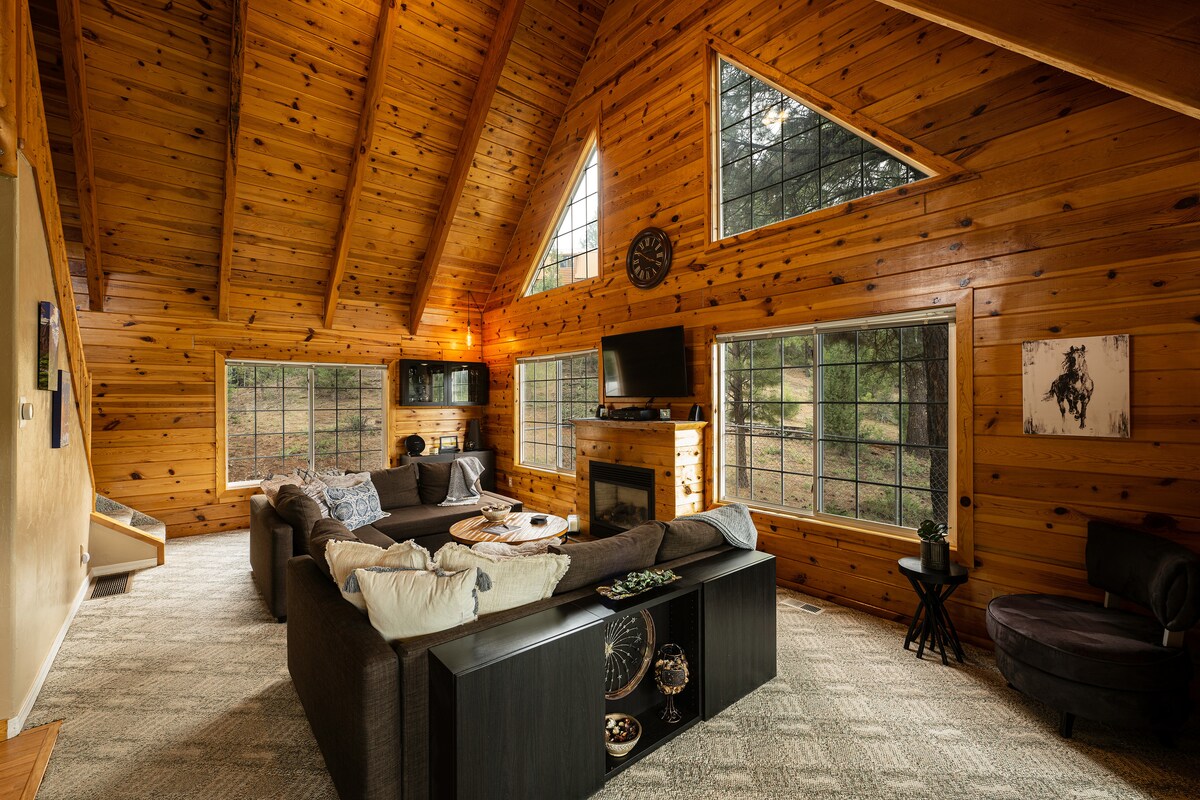 The living room features a high, vaulted ceiling with exposed wood beams. Large triangular windows flood the space with natural light, framing views of the surrounding trees. A cozy sectional sofa and a circular lounge chair provide seating, while a fireplace is situated beneath a mounted television.
