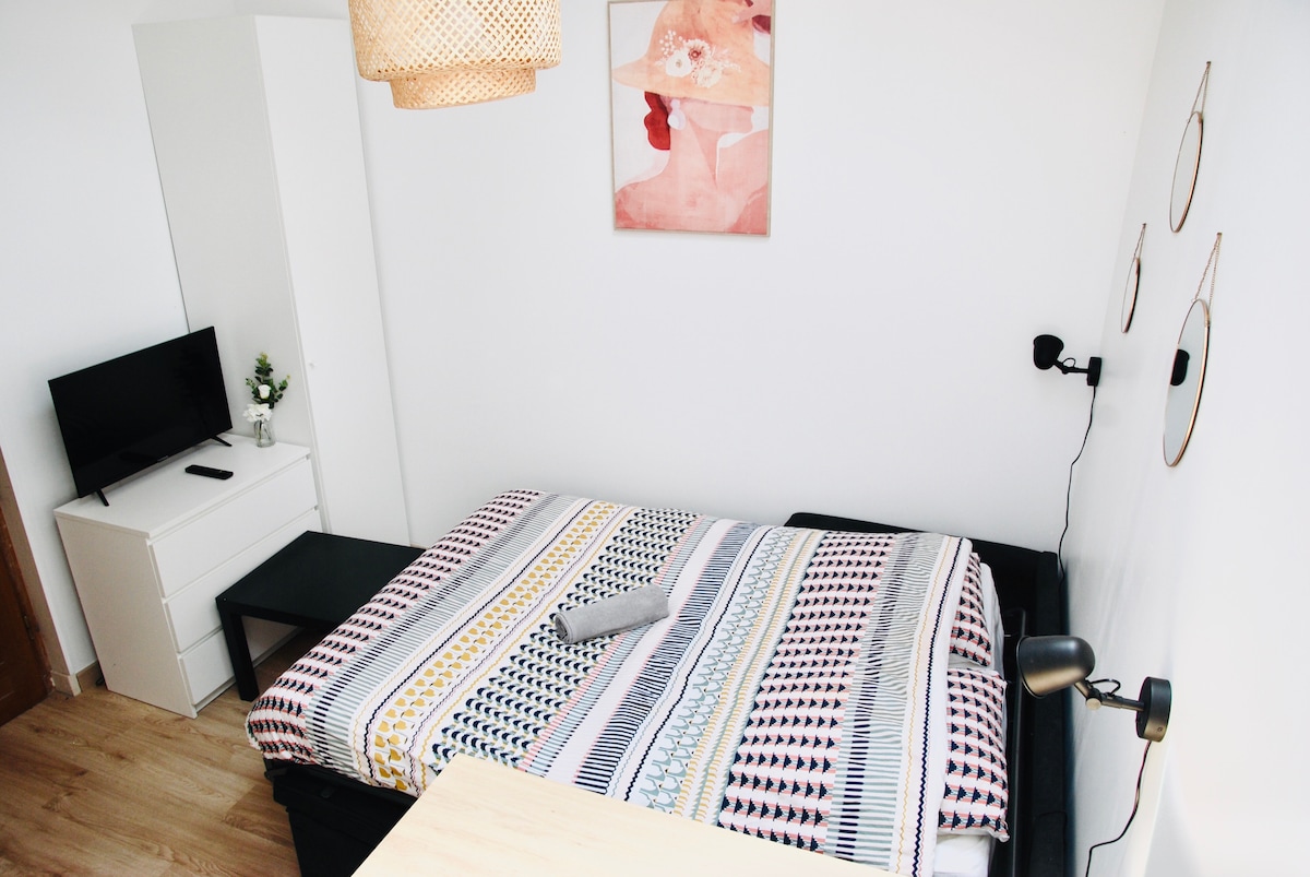 The sleeping area features a spacious bed adorned with a patterned blanket. A small television is positioned on a white dresser, and a wooden table is nearby. Wall decorations include a framed artwork and three circular mirrors. Natural light fills the room from a window.