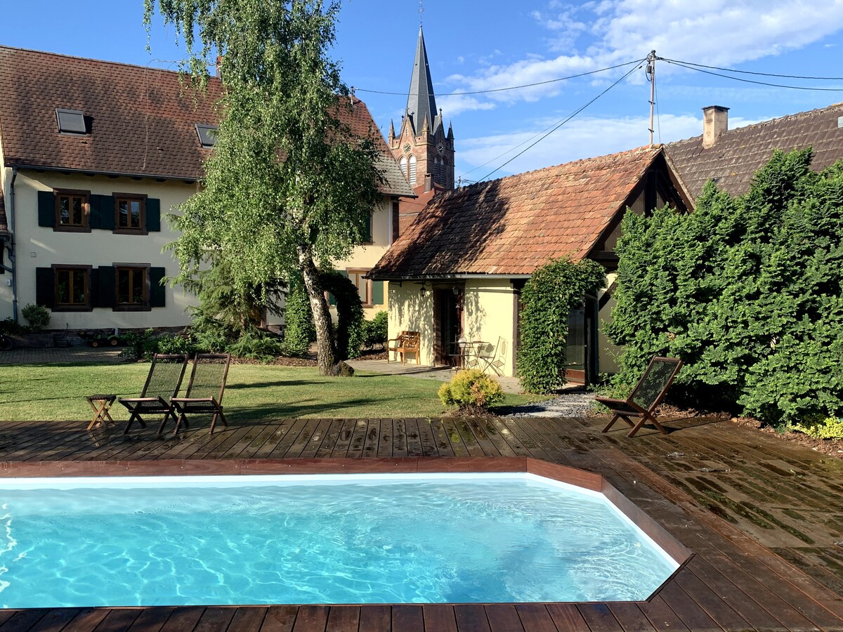 A serene outdoor space features a wooden deck surrounding a heated pool. Lush greenery and neatly trimmed hedges provide a sense of privacy. In the background, a well-maintained garden is visible, along with charming houses and a church spire reaching towards the sky.