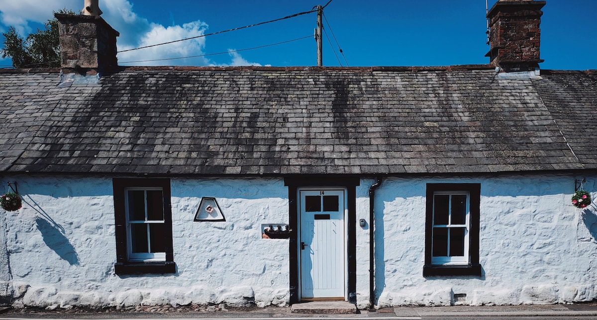 The exterior of a charming cottage is showcased, featuring whitewashed stone walls and a slate roof. A central door is framed by decorative elements, with two windows on either side. Lush greenery is visible in the background under a clear blue sky.