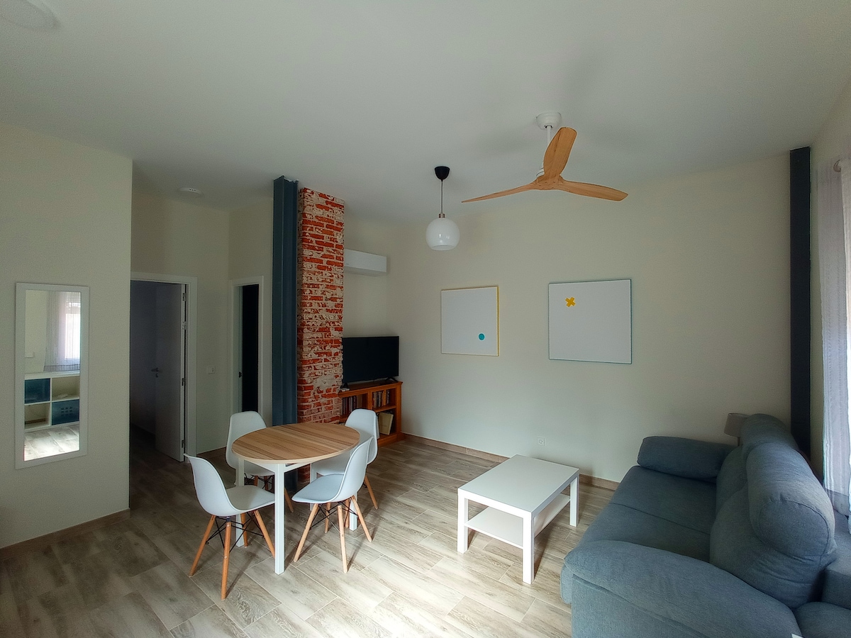 A spacious living area is defined by light wooden flooring and neutral-colored walls. A comfortable gray sofa faces a modern TV setup, with a round dining table surrounded by sleek white chairs. A decorative ceiling fan is visible, enhancing the airy ambiance.