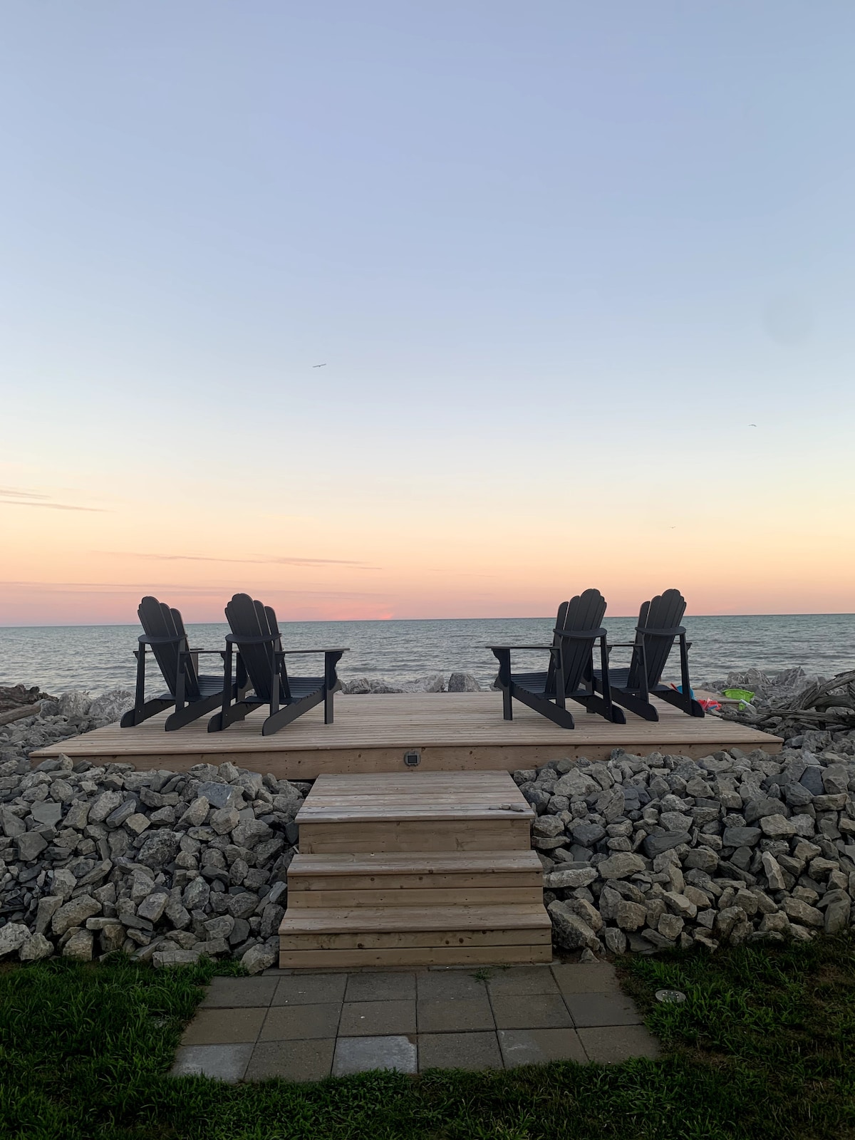 A wooden dock built on a stone break wall overlooks the water, featuring four Adirondack chairs arranged for seating. A soft gradient of colors in the sky reflects the sunset, with the calm waters of Lake Erie in the background.