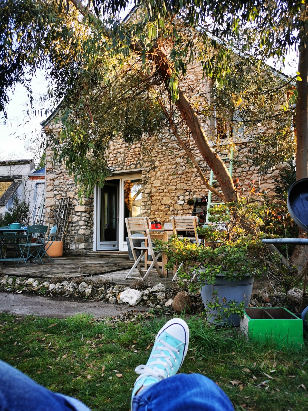 The image shows a charming stone house surrounded by greenery. Outdoor seating areas are visible, including several chairs and a small table on a wooden deck. A tree partially shades the area, adding to the natural ambiance of the garden.