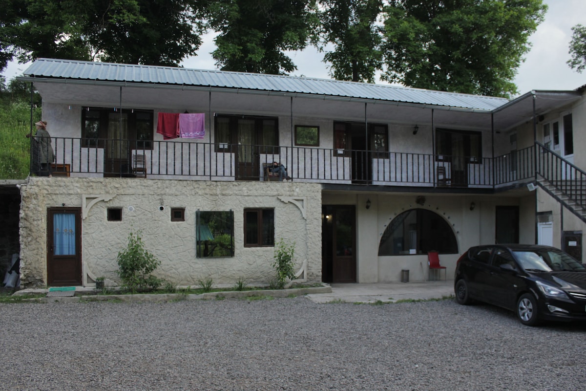 A two-story guesthouse is depicted with a light-colored exterior and a metal roof. Several doors lead to individual rooms, with a balcony overhead. A gravel parking area is shown in the foreground, alongside modest landscaping and a parked car.