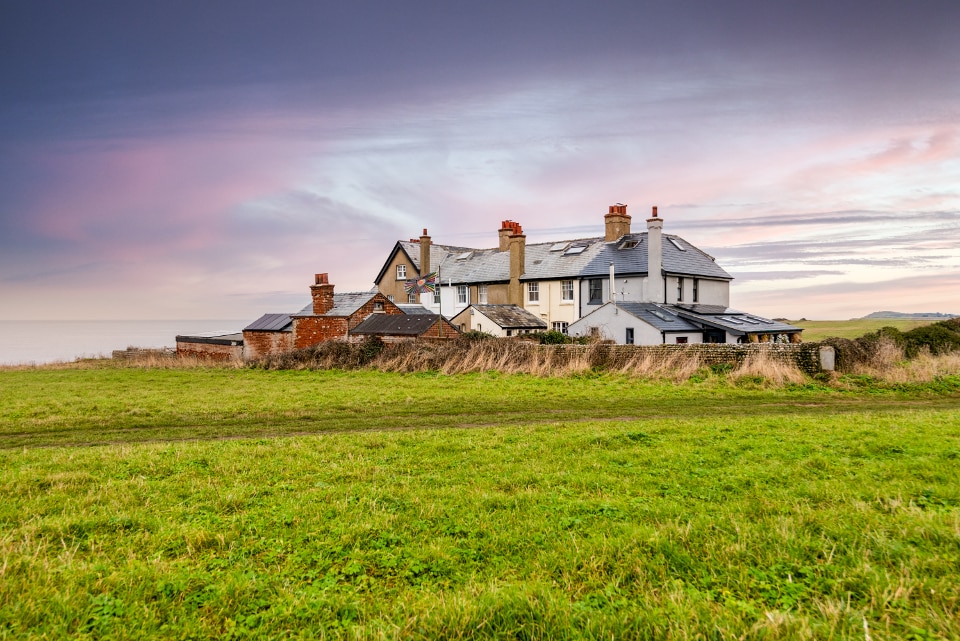 The cottage, set against a colorful sunset sky, features traditional architecture with a series of brick chimneys. Surrounding grassy fields lead up to the building, providing a natural buffer. The structure is positioned near the coastline, with the sea visible in the background.