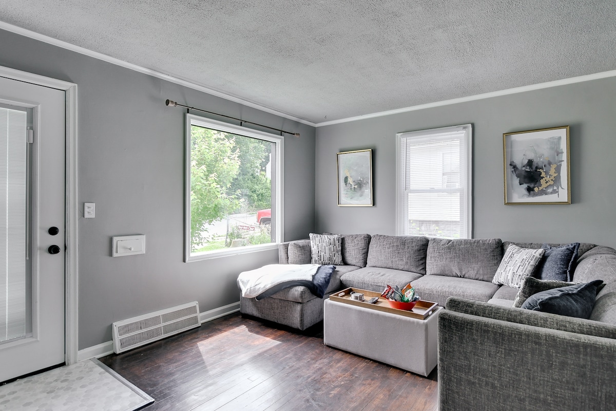 A cozy living room is highlighted by a large gray sectional sofa and a textured coffee table. Natural light enters through a sizable window, revealing views of the outdoors. Two framed art pieces adorn the walls, complementing the neutral color scheme.