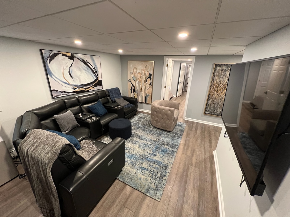A modern living area features a black sectional sofa and an upholstered chair set against a light grey wall. Artwork adorns the walls, adding visual interest. A large television is mounted on one wall, and ambient lighting brightens the space.