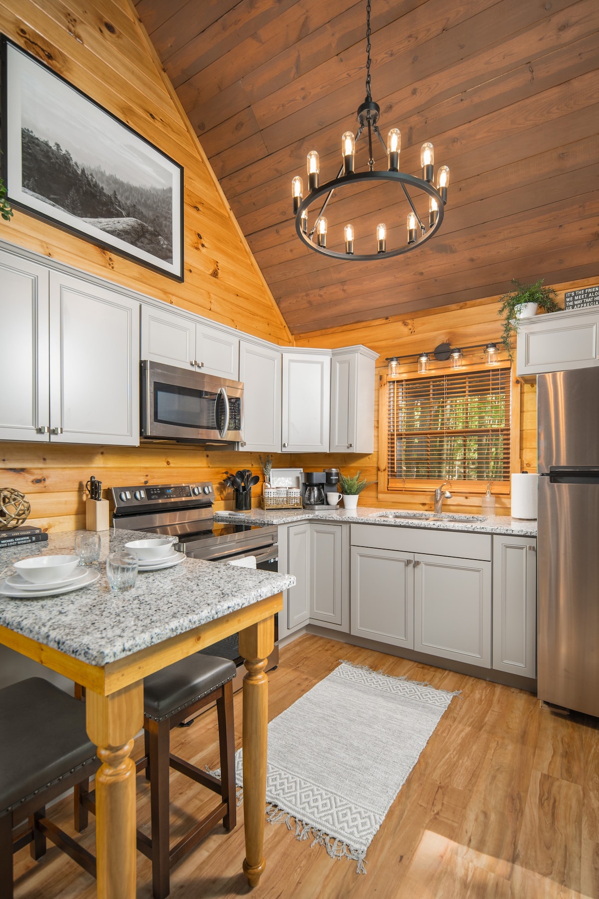 The kitchen features modern appliances, including a microwave and a stainless-steel refrigerator. A granite countertop is complemented by wooden cabinetry and a rustic light fixture. A small dining area with two chairs is set beside the window, allowing natural light to enhance the warm wood tones.