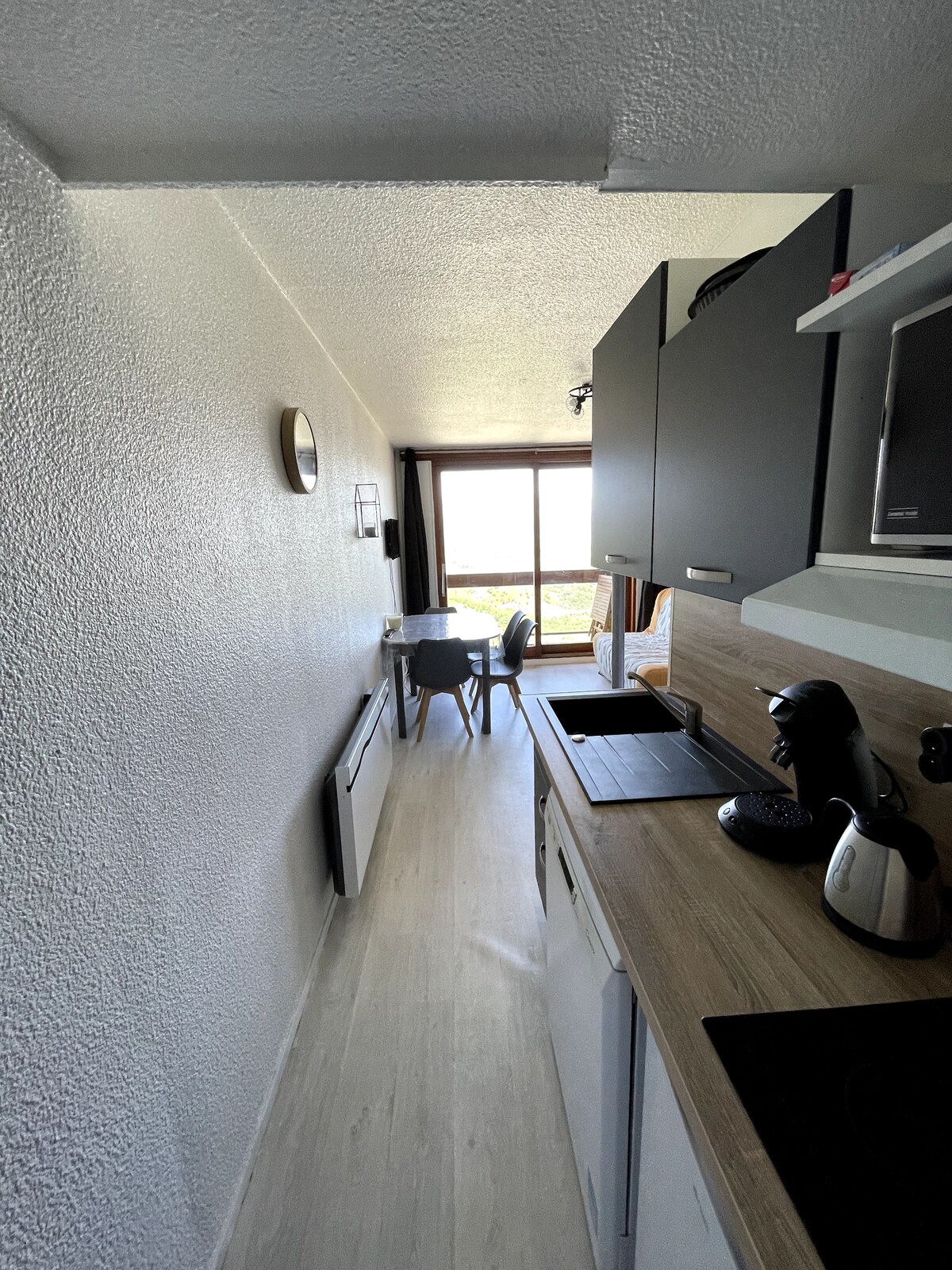 A modern kitchenette is visible, featuring dark cabinetry and light wood countertops. A coffee maker and kettle are positioned on the counter. A dining table for four, with a view through a large window, enhances the space. Natural light illuminates the area.