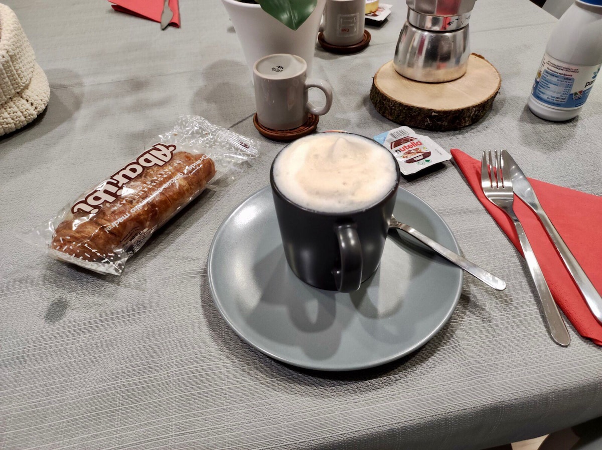A grey plate holds a cup of cappuccino, topped with frothy foam, next to a wrapped pastry. A small ceramic cup sits behind, alongside a wooden coaster and a pack of sugar. A knife and fork are neatly placed on a table covered with a light grey tablecloth.
