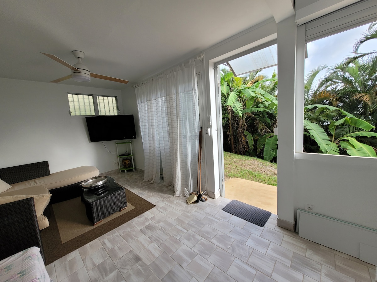 A spacious living area features a comfortable seating arrangement and a wall-mounted television. Bright natural light enters through large windows with sheer curtains, revealing views of a lush green outdoor space with tropical plants. A ceiling fan is overhead, enhancing air circulation.