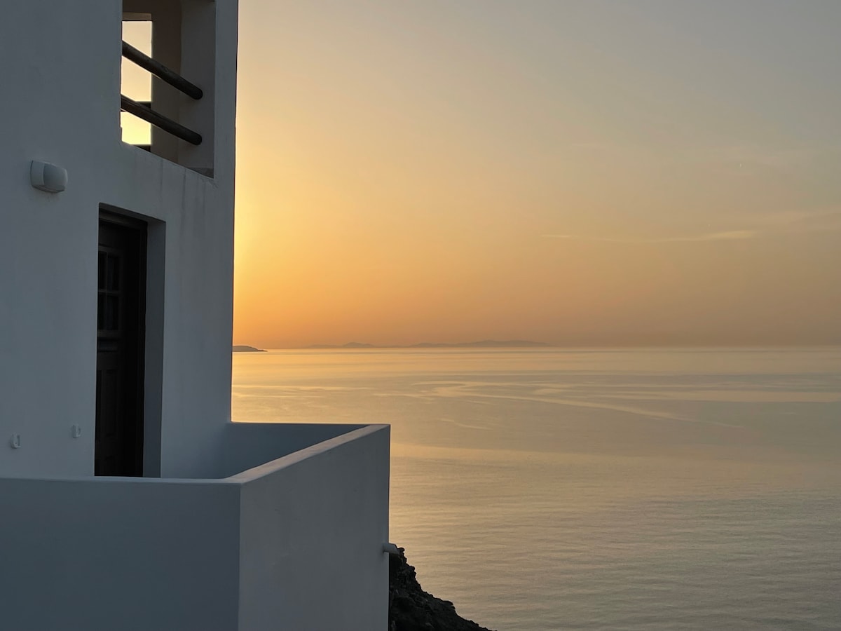 A serene sunset view is captured from the balcony of a white-walled building, with soft orange and yellow hues reflecting on the calm sea. The faint outline of distant islands can be seen on the horizon, creating a tranquil atmosphere.