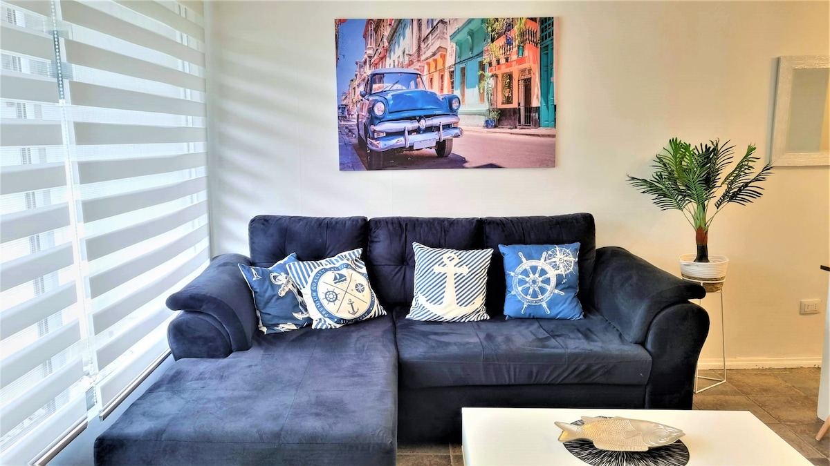 A spacious living area features a dark couch with decorative pillows in various shades of blue, complementing a vibrant wall art piece depicting a classic car. A light-colored coffee table sits in front, and a small potted plant adds a touch of greenery.