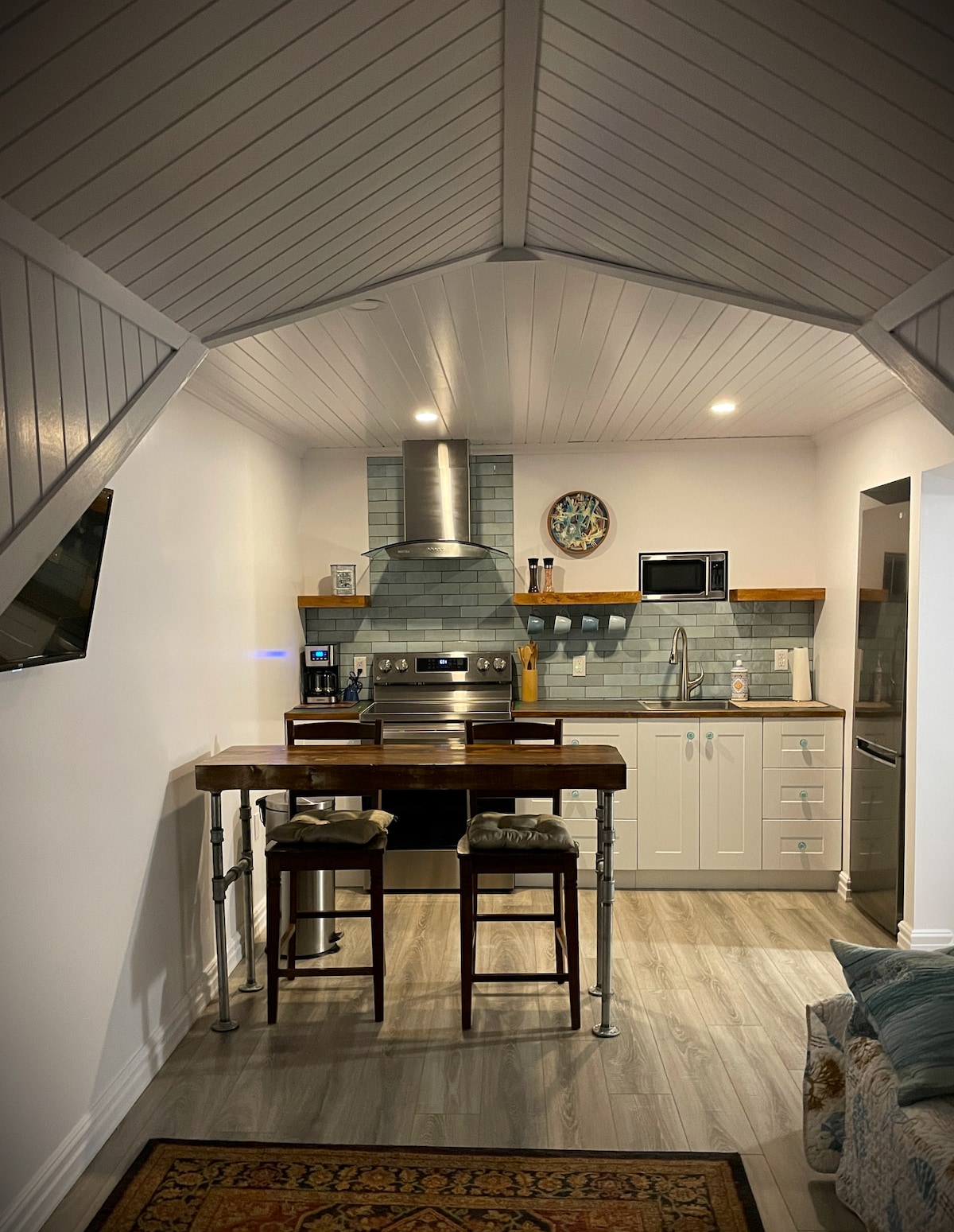 The kitchen area features modern appliances, including a stainless-steel stove, microwave, and refrigerator. A wooden counter with two stools offers a comfortable dining space. Subtle grey tiles accent the walls, while the vaulted ceiling enhances the open and airy feeling of the space.