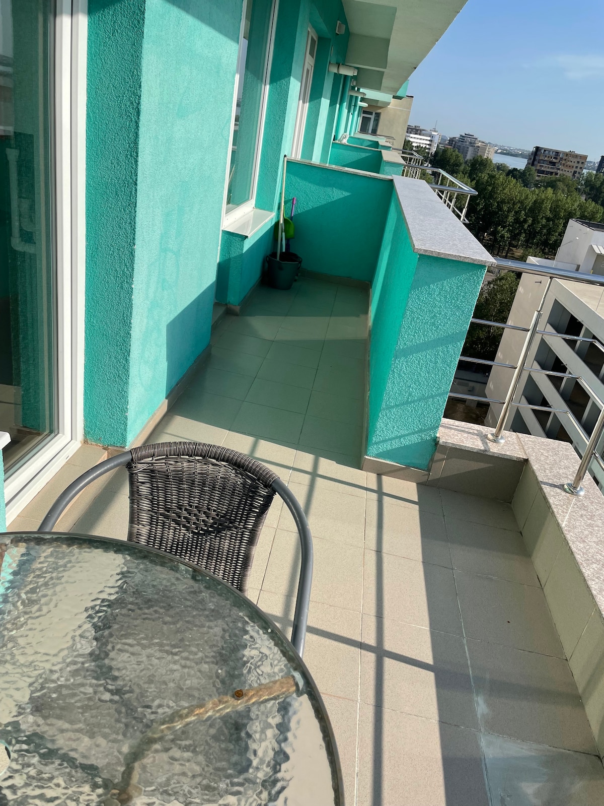 A balcony area features a glass table with a single wicker chair, surrounded by vibrant turquoise walls. Natural light illuminates the space, enhancing views of the surrounding buildings and distant sea. The flooring is tiled, providing a clean and open feel.