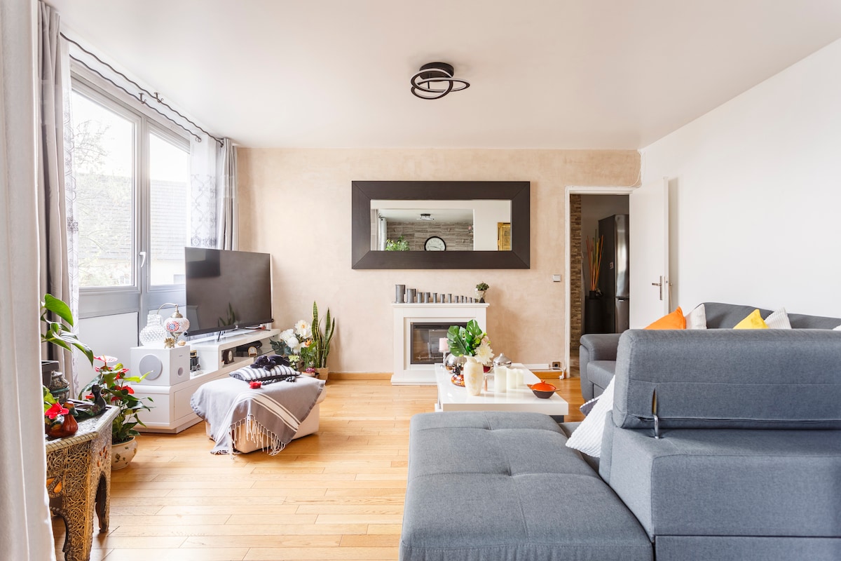 A spacious living room is illuminated by natural light from large windows. Gray sofas are arranged around a coffee table, while a flat-screen TV is positioned near a decorative mirror. Potted plants and decor add touches of greenery and warmth to the neutral-toned space.