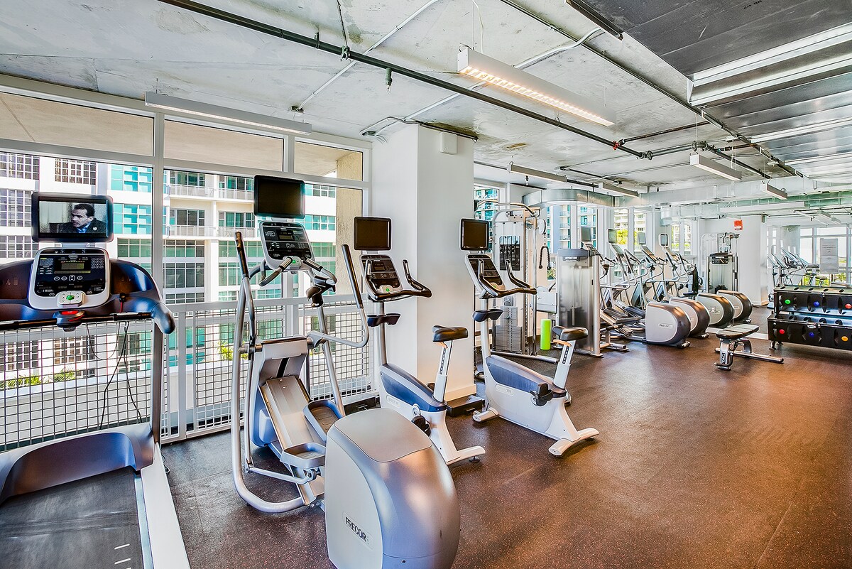 A modern gym space is equipped with various fitness machines, including treadmills and elliptical trainers. Bright natural light filters in through large windows, illuminating the space. Free weights and additional workout equipment are organized along one wall, creating an inviting atmosphere for exercise.