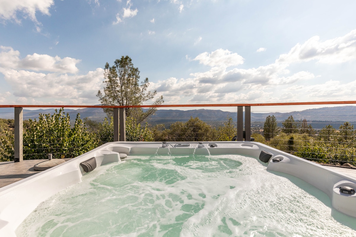 A spacious hot tub is positioned on a deck, offering expansive views of the surrounding landscape. The bubbling water reflects the sky, with clouds drifting overhead. Lush greenery lines the area, enhancing the serene outdoor setting.