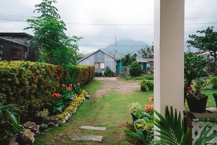 A well-maintained garden pathway leads through lush greenery and vibrant flower beds, framed by colorful plants. In the background, modest structures can be seen, set against a backdrop of rolling hills and cloudy skies.