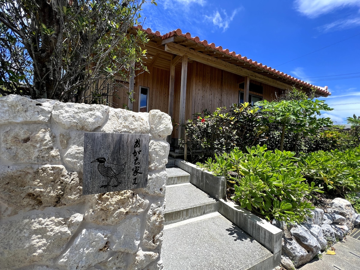 A rustic entrance is framed by lush greenery leading to a wooden building with a red-tiled roof. A stone path and steps guide the way, while a decorative sign features an artistic depiction of a bird, welcoming guests.