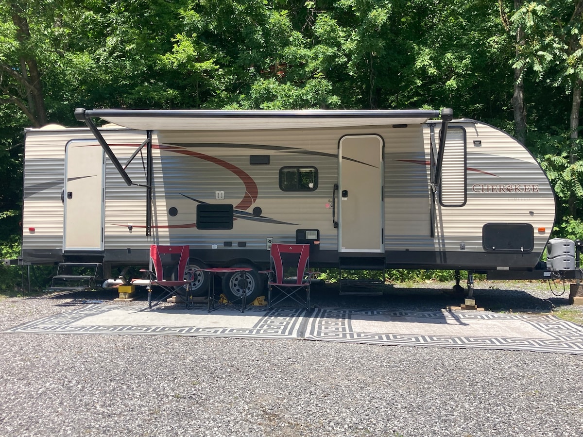 A spacious camper is positioned on a gravel area, featuring a retractable awning for shade. Two red chairs are placed on an outdoor rug, creating a cozy seating arrangement. Lush green trees provide a natural backdrop, enhancing the outdoor experience.