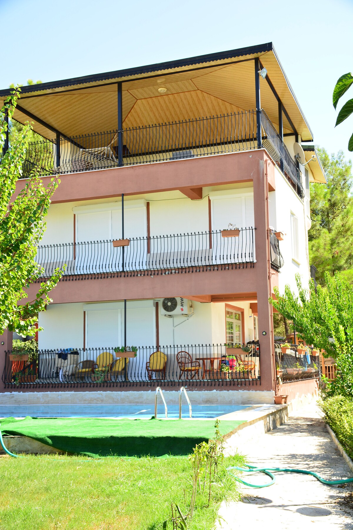 A three-story residence is depicted with a terrace featuring ample seating arrangements. The ground level includes a pool shaded by nearby trees. Large windows bring natural light into the structure, complemented by greenery surrounding the property.