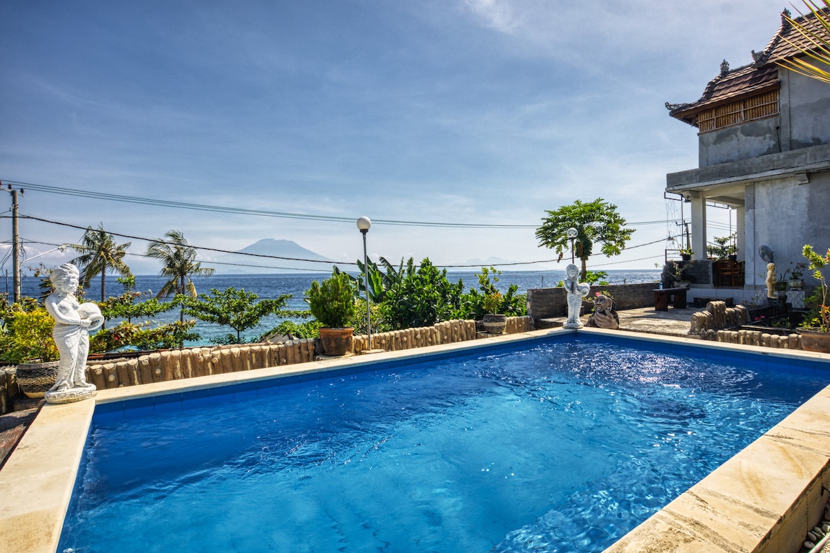 A clear swimming pool is situated within a stone patio, offering views of the ocean. Lush greenery surrounds the area, with decorative sculptures enhancing the landscape. The distant mountains are visible under a clear blue sky, creating a serene atmosphere.