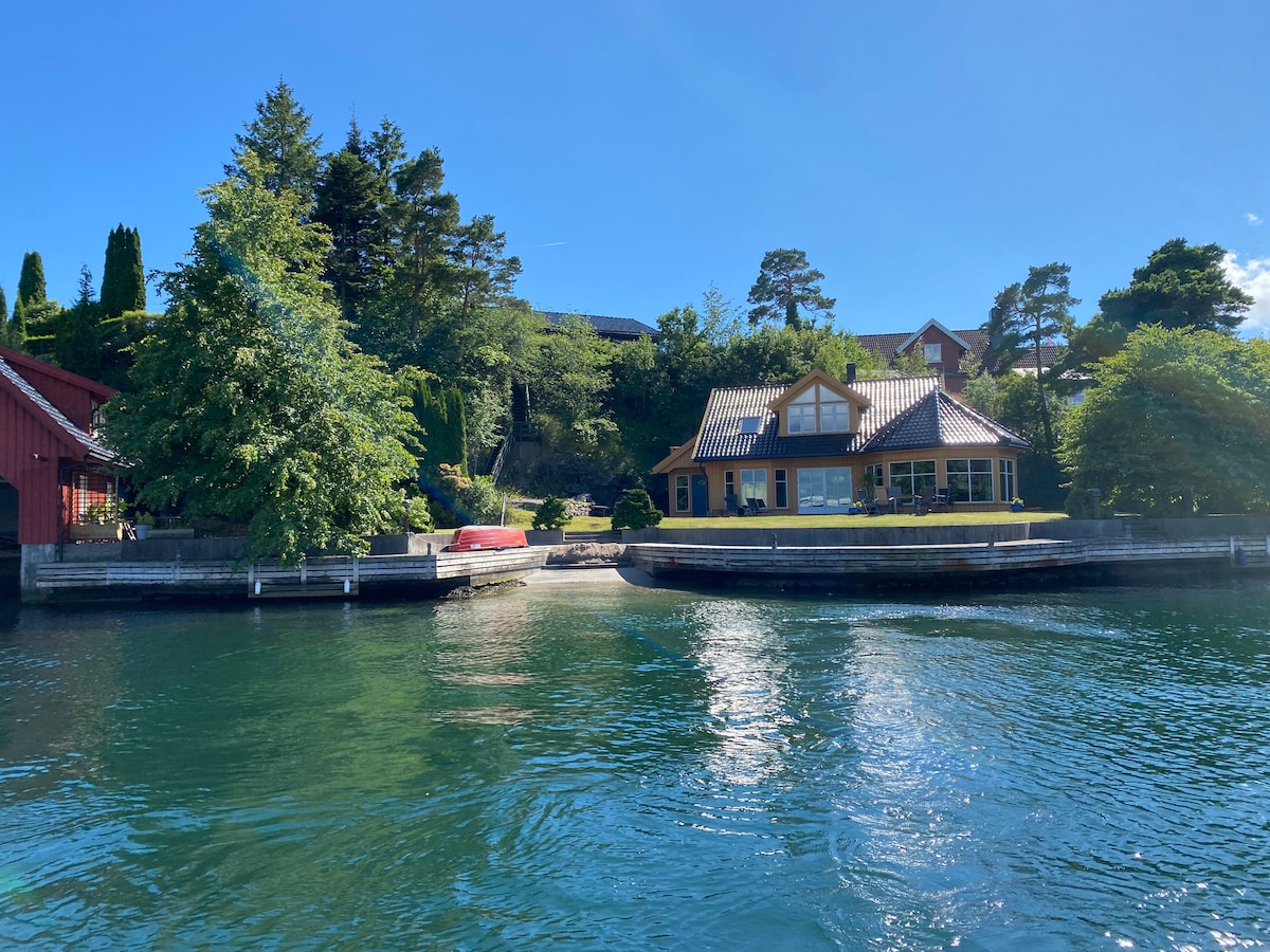 A waterfront property is surrounded by lush greenery, showcasing a modern design with large glass windows. A private dock is visible alongside the serene water, while trees create a natural border to the scene. The bright blue sky adds to the tranquil setting.