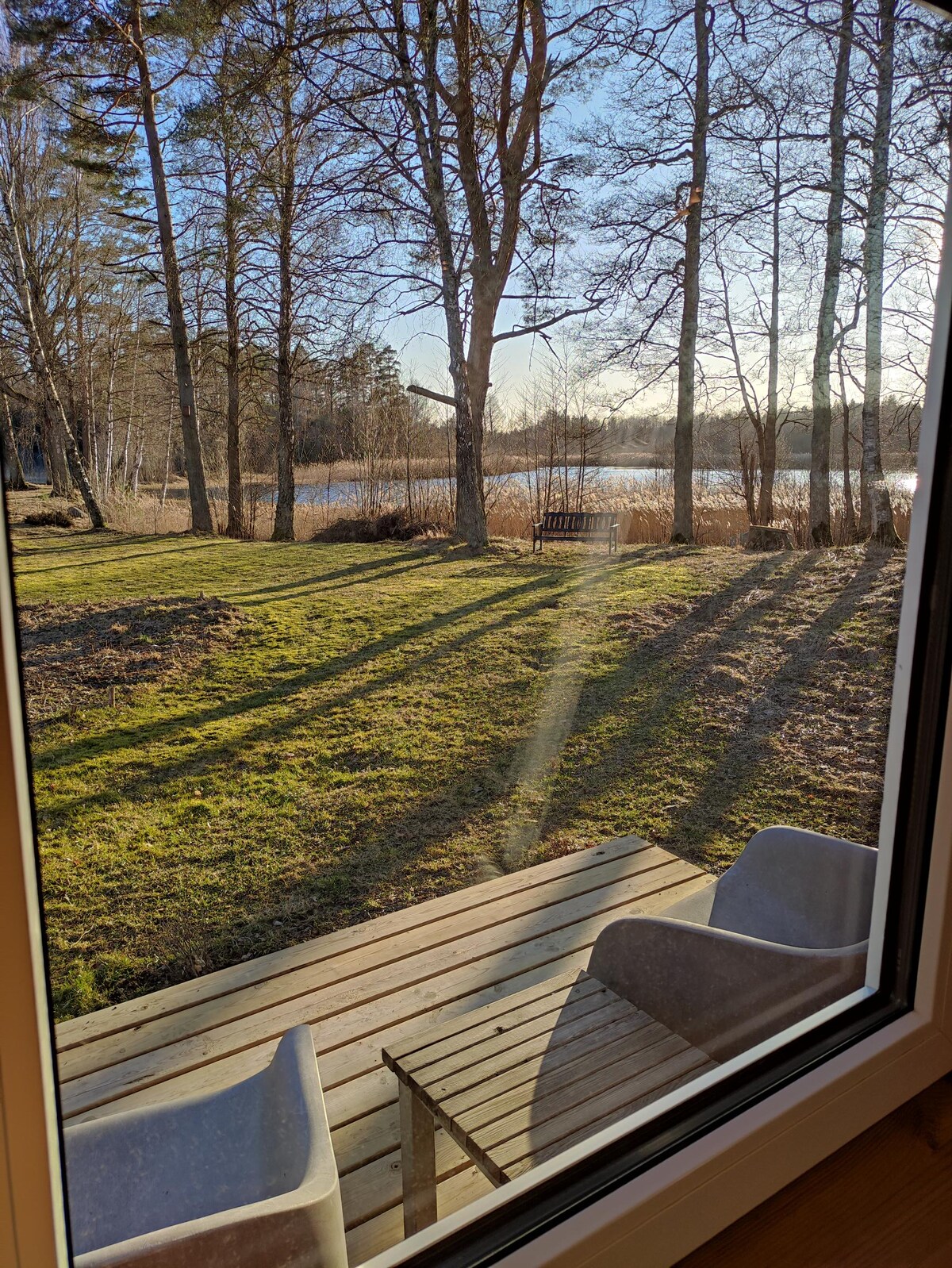 The view from the window reveals a grassy area with scattered trees, leading to a serene lakeside. Two white chairs are positioned on a wooden terrace, inviting relaxation while overlooking the water, which reflects the sunlight.