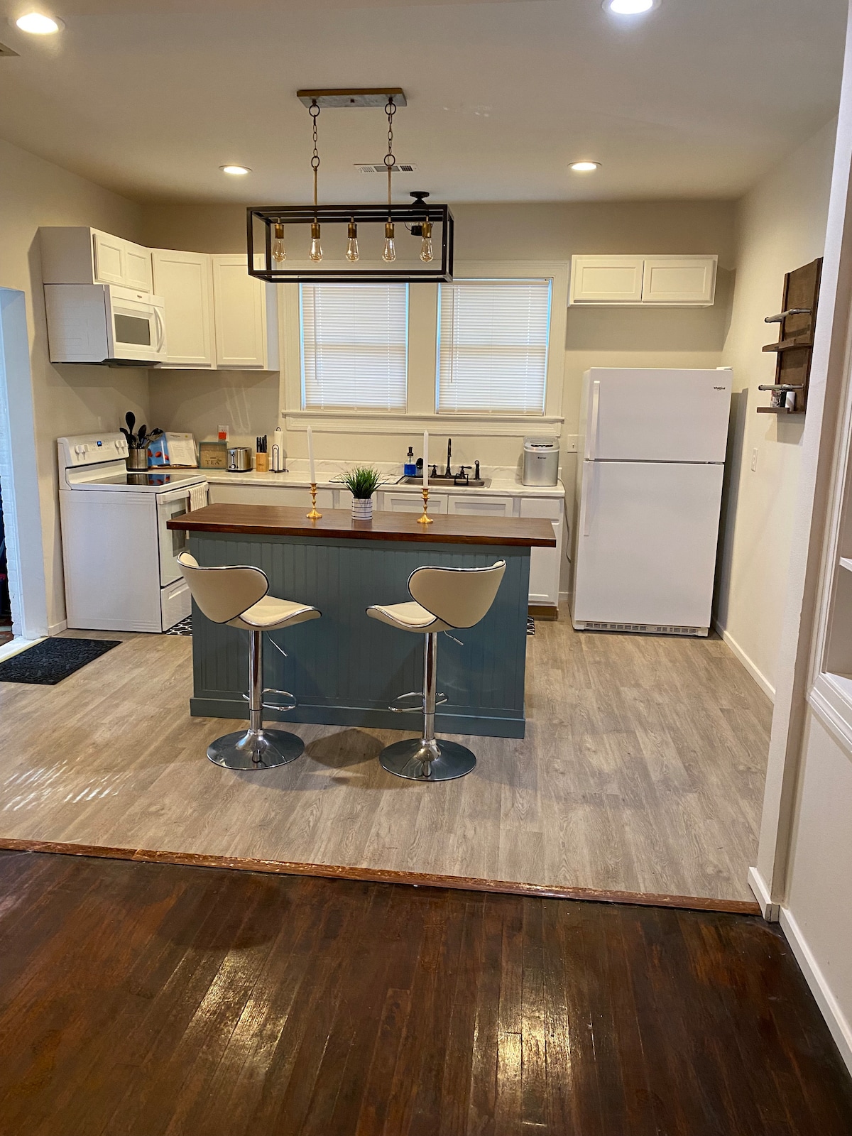 The kitchen features modern cabinetry with a central island and two sleek stools. Appliances include a microwave and refrigerator, while light streams in through the windows above the sink, highlighting the spacious layout.