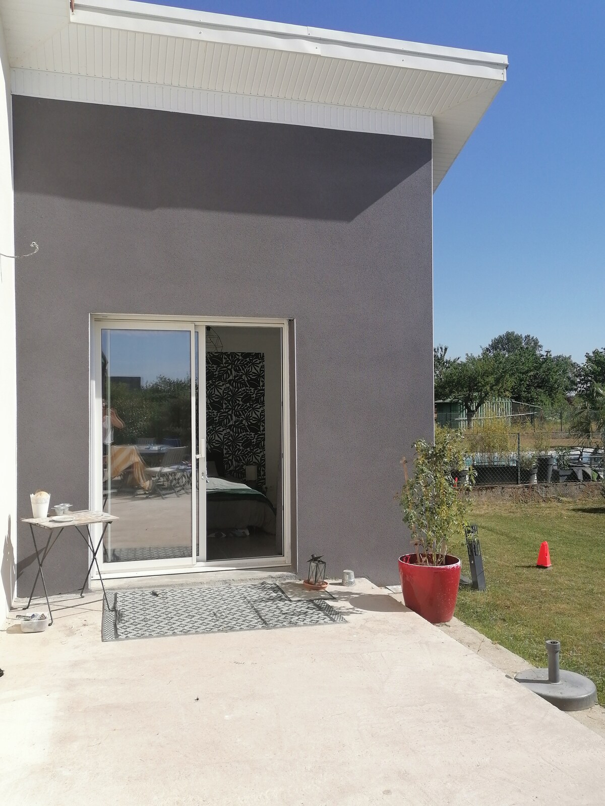 A modern exterior of a small independent studio is visible, featuring a sliding glass door that leads to the interior. A small outdoor table and a potted plant add a touch of greenery beside the door, with a well-maintained lawn stretching into the background.