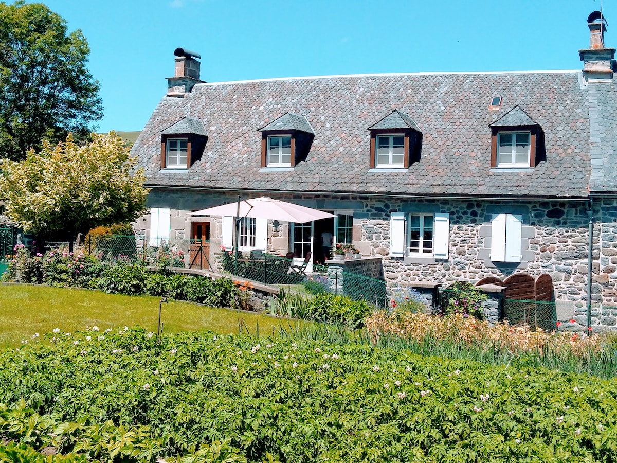 The exterior of a stone house features a sloped roof with multiple dormer windows. A large terrace with an awning is visible, surrounded by well-maintained gardens filled with greenery and flowers. Natural light brightens the scene under a clear blue sky.