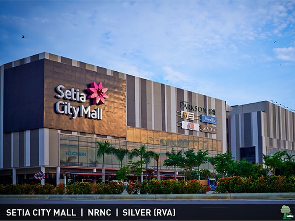 The exterior of Setia City Mall is displayed, featuring a modern facade with varied textures and colors. The iconic mall logo is visible, along with signage for various retailers. Lush greenery surrounds the entrance, contributing to the vibrant atmosphere of the shopping area.