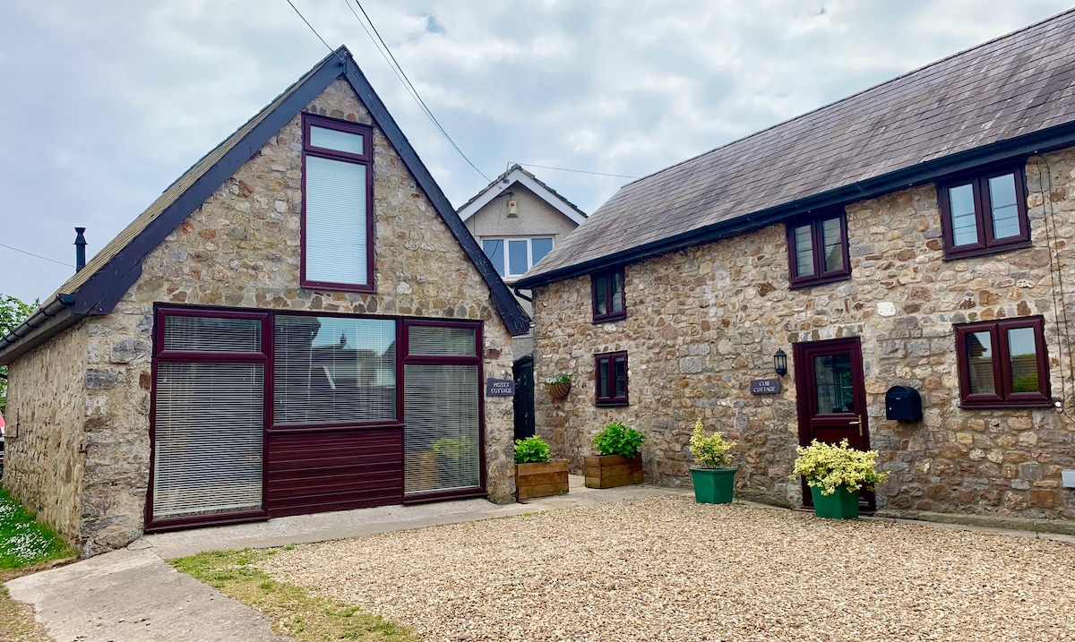The exterior of a stone cottage is highlighted, featuring a combination of large windows and traditional elements. A gravel driveway leads to the entrance, surrounded by greenery in planters. The rustic charm of the structure is enhanced by its inviting layout against a partly cloudy sky.