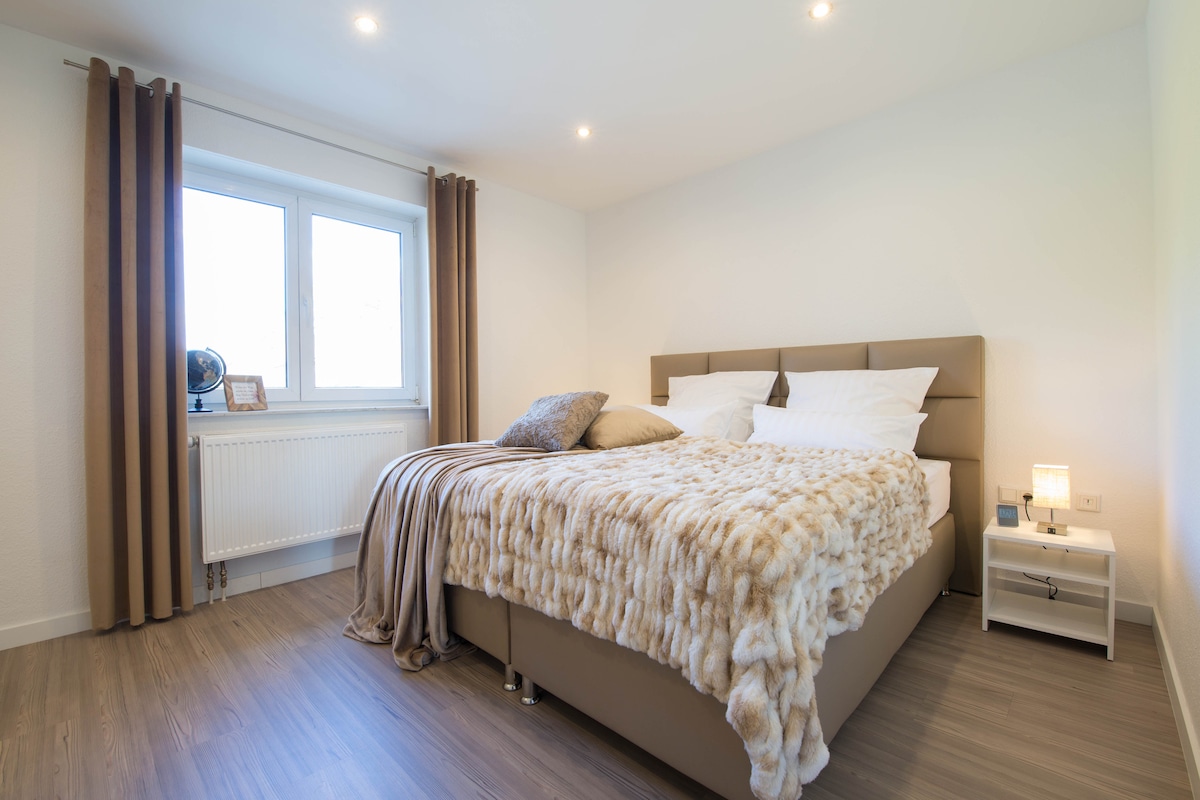 A modern bedroom features a spacious bed dressed in soft linens and a textured throw blanket. Natural light enters through a large window, complemented by neatly arranged curtains. A bedside table holds a minimalist lamp, enhancing the room's calm and organized atmosphere.