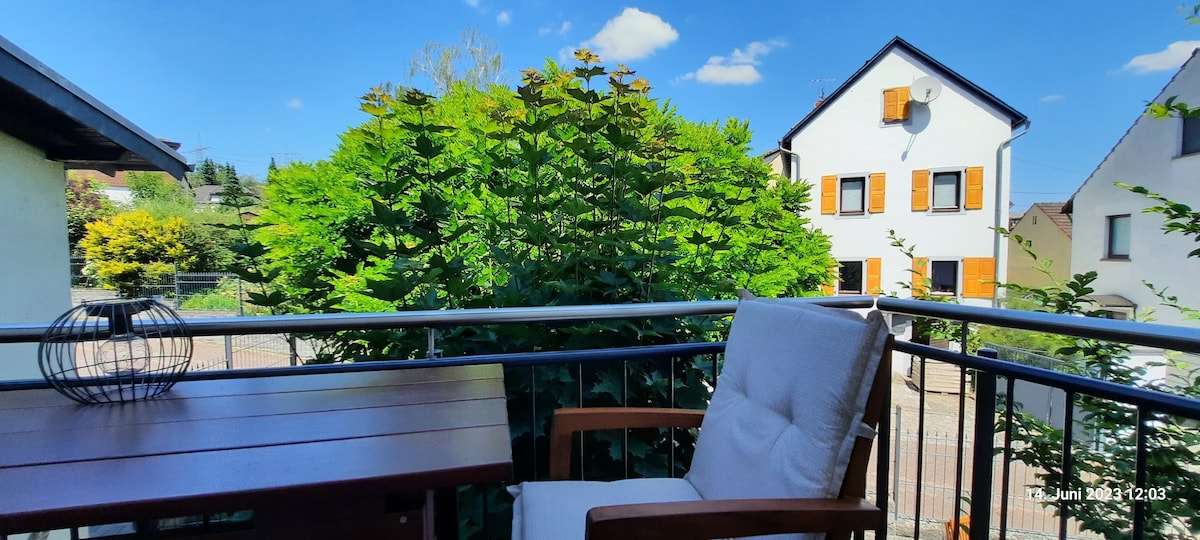 A private balcony with a wooden table and a comfortable chair offers views of lush greenery. The surrounding foliage creates a serene atmosphere, while a bright blue sky and fluffy clouds are visible in the background, enhancing the outdoor experience.