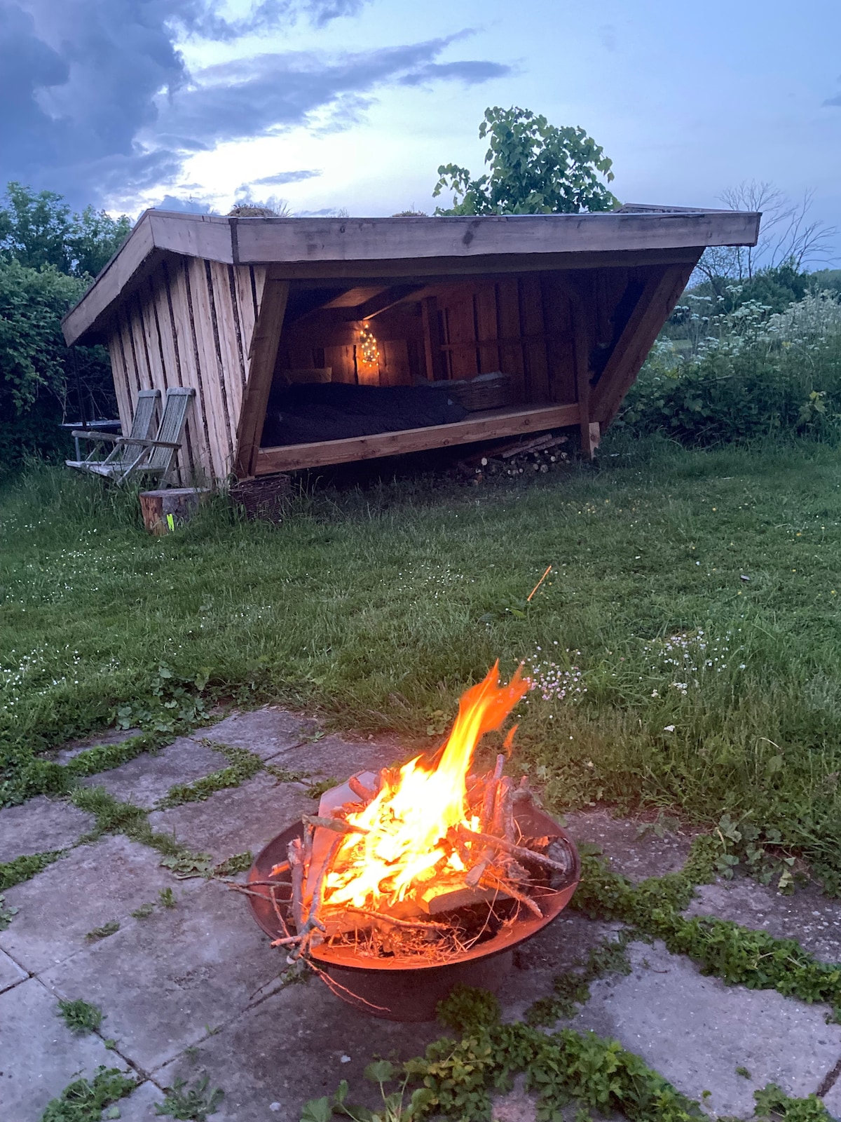 A sheltered structure sits on a grassy area, featuring an angled roof and large overhanging eaves. A cozy outdoor fire pit is positioned nearby, with flames flickering against the evening sky. Surrounding greenery adds to the natural setting, creating a serene ambiance.