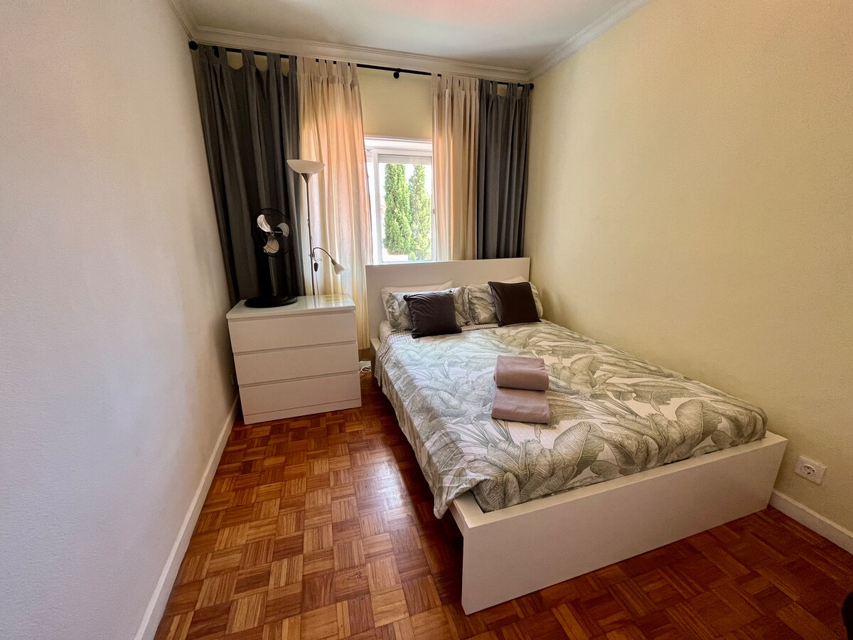 A bright double room features a spacious bed with a leafy patterned duvet and two pillows. Natural light enters through a window, flanked by light and dark curtains. A bedside lamp stands next to the bed, and a chest of drawers is positioned against the wall.