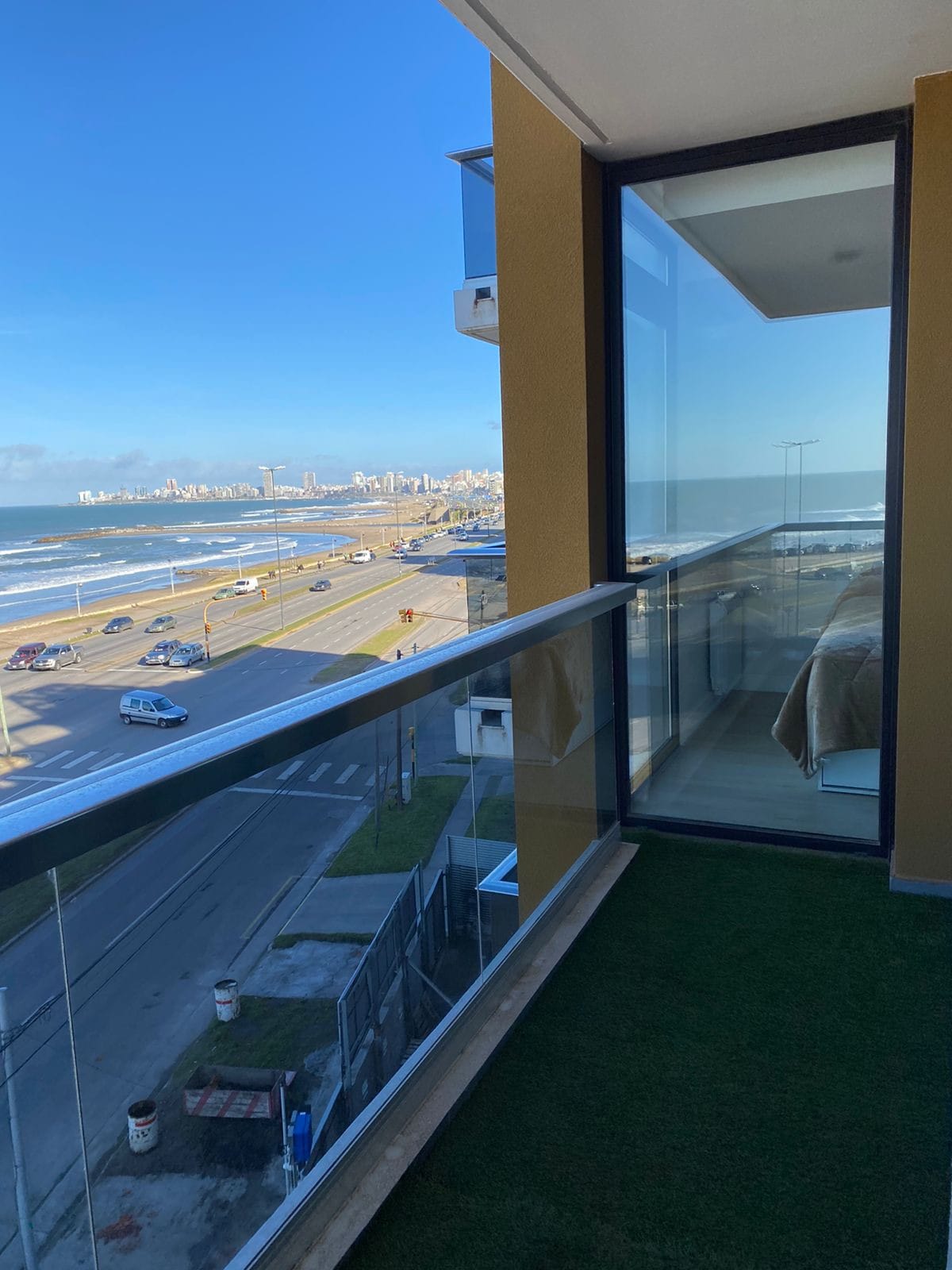 A spacious balcony overlooks the serene ocean, offering unobstructed views of the coastline and city skyline. Glass railings provide safety while allowing for clear sightlines. The bright blue sky complements the tranquil sea, with urban elements visible in the distance.